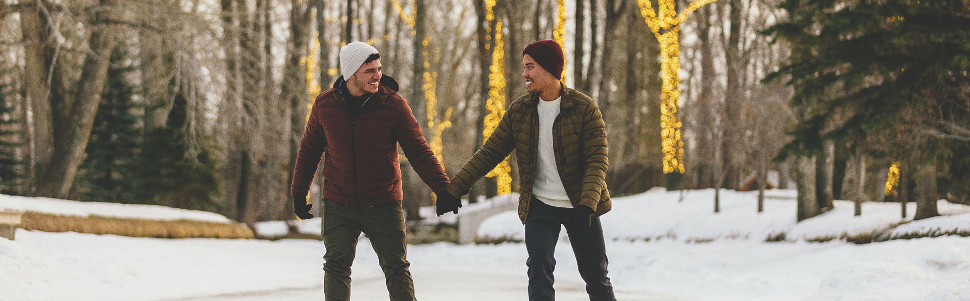 LGBTQ couple skating through Bowness Lagoon holding hands with lit up trees behind them