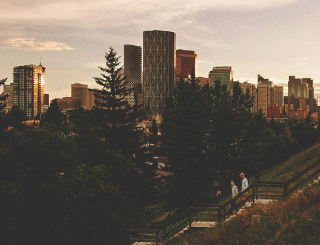 two friends walk down an outdoor staircase lined with trees. the downtown calgary skyline is prominent in the background.