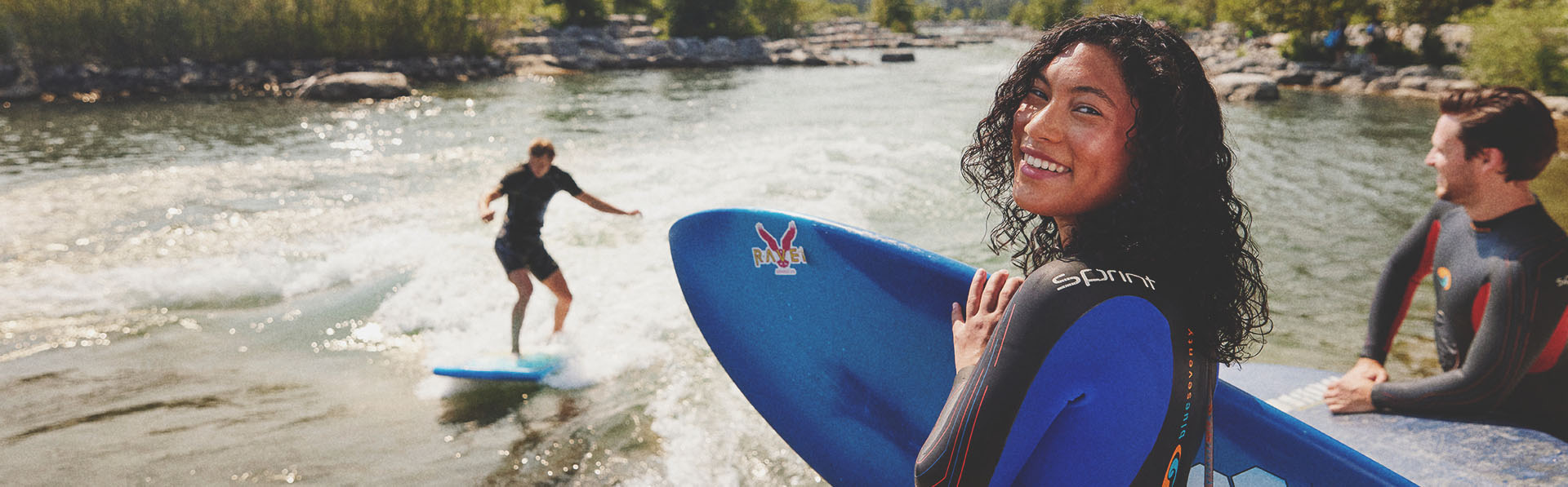 woman looks toward the camera holding a surfboard as a friend surfs Harvie Passage on the Bow River in the backgrounds