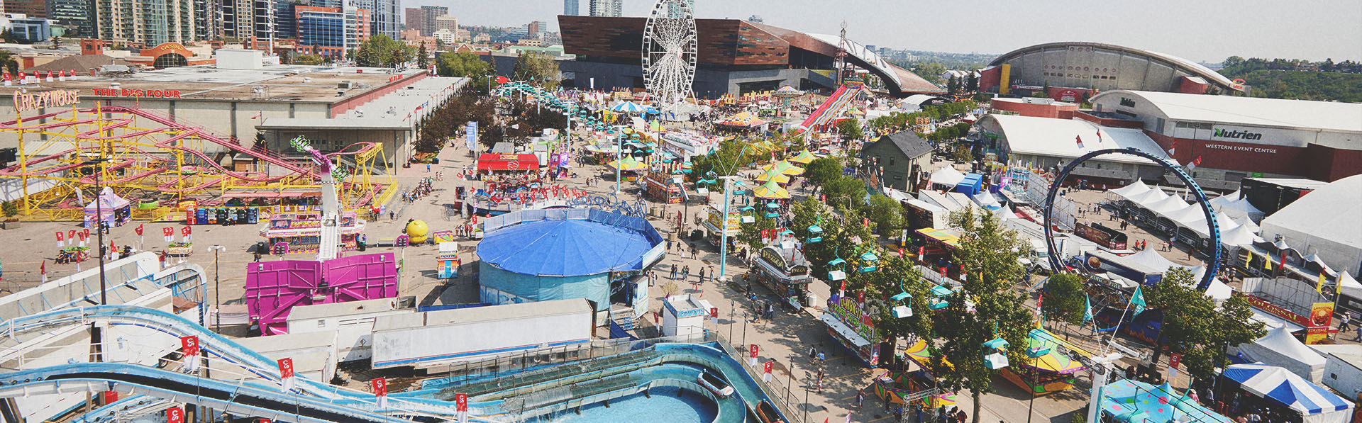 Aerial view of the Calgary Stampede grounds featuring the Saddledome, midway rides, and crowds of visitors.