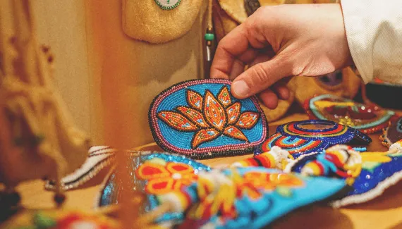 Someone picks up a Beaded Orange Flower Barrette from a shelf full of other beadwork arts and crafts as well as ceremonial and medicine bags
