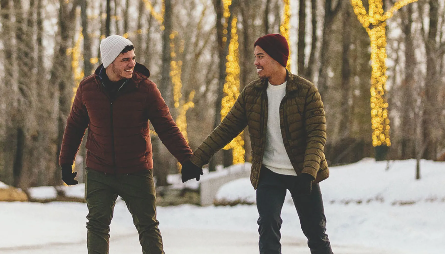 LGBTQ couple skating through Bowness Lagoon holding hands with lit up trees behind them