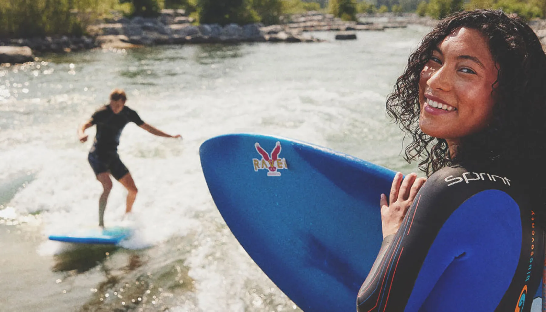 woman looks toward the camera holding a surfboard as a friend surfs Harvie Passage on the Bow River in the backgrounds