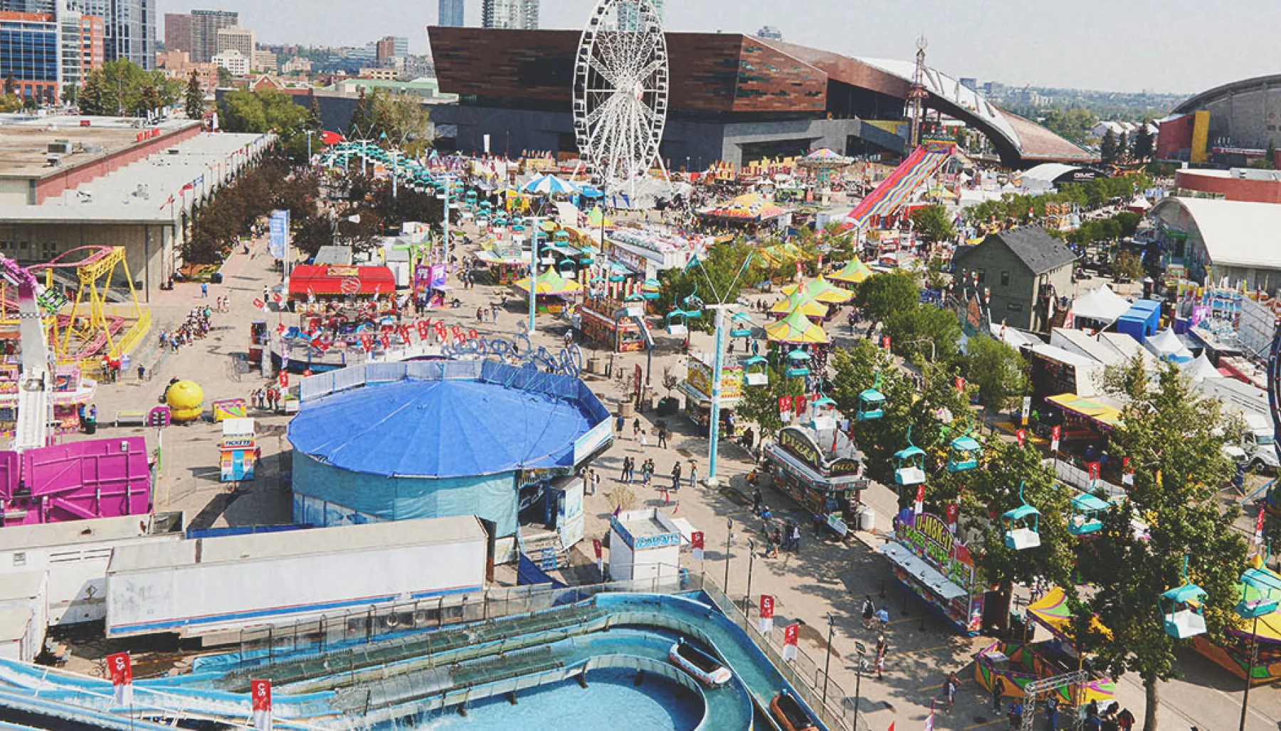 Aerial view of the Calgary Stampede grounds featuring the Saddledome, midway rides, and crowds of visitors.