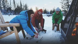 three friends lacing up skates beside a outdoor firepit at the Bowness lagoon skating rink