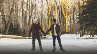 LGBTQ couple skating through Bowness Lagoon holding hands with lit up trees behind them