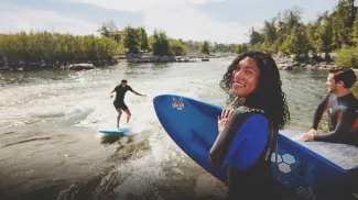 woman looks toward the camera holding a surfboard as a friend surfs Harvie Passage on the Bow River in the backgrounds