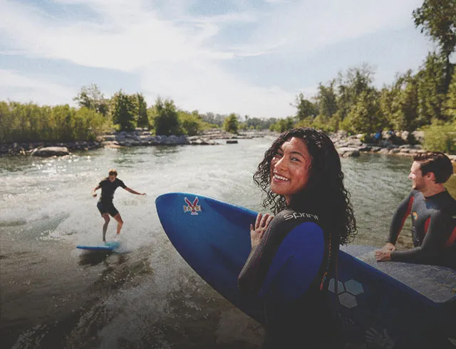 woman looks toward the camera holding a surfboard as a friend surfs Harvie Passage on the Bow River in the backgrounds