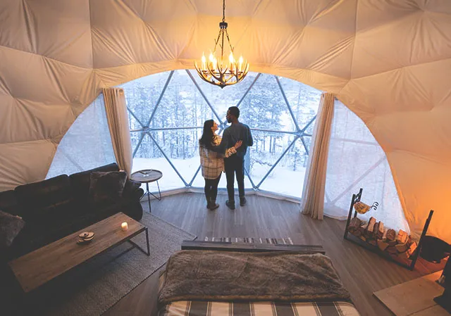 Couple stands inside a luxury camping dome looking outside during winter