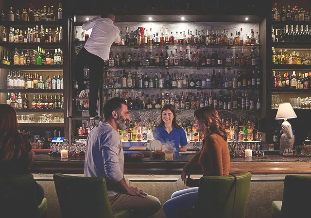 couple sitting at a bartop as the server reveals a smoked cocktail. A bartender climbs a ladder to reach a top shelf bottle.