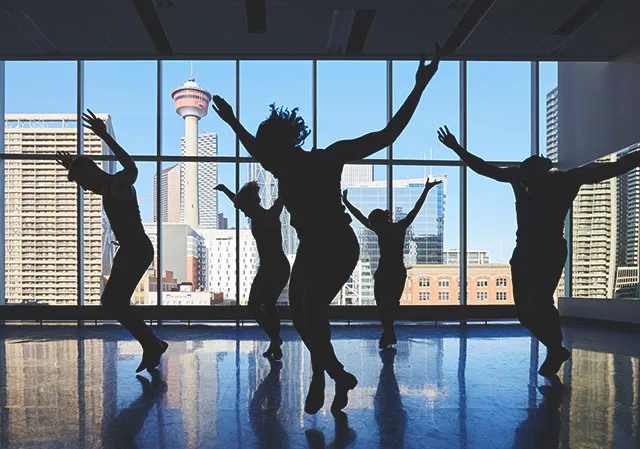 silhouette of DJD dancers performing in their studio with the Calgary skyline and tower in the background through the windows