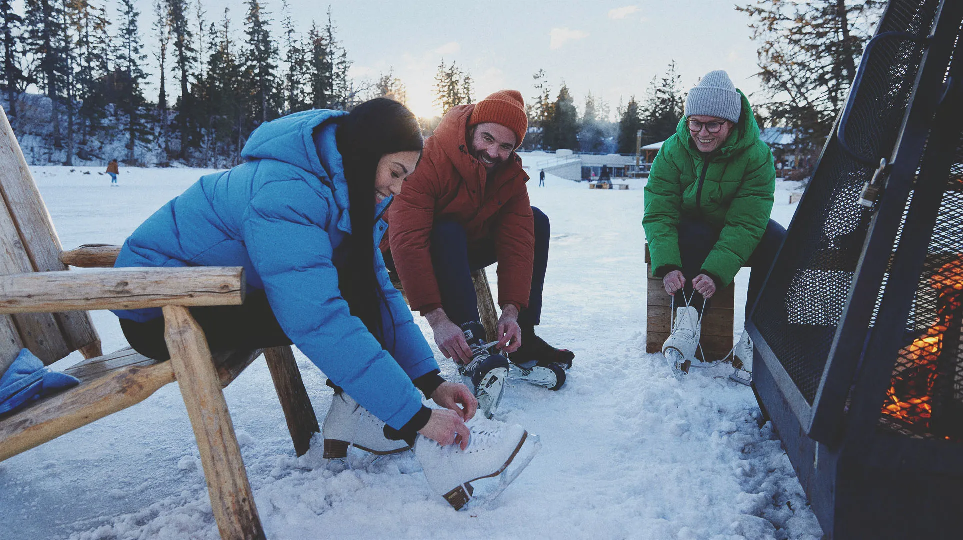 three friends lacing up skates beside a outdoor firepit at the Bowness lagoon skating rink
