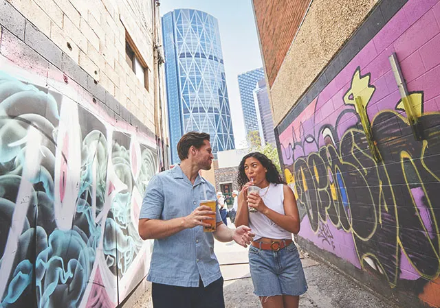couple sipping bubble tea and walking through mural alley in Calgary's Chinatown, with the Bow tower behind them peeking between two buildings