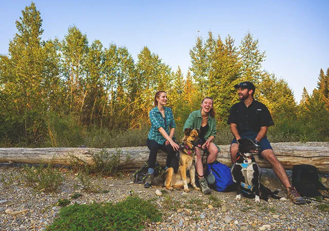 group of three friends taking a break from hiking in Fish Creek Park to sit on a log with their dog